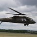 A United States Army UH-60 Black Hawk helicopter, tail number 908, is captured in low-altitude flight over what appears to be an airfield or military installation. The helicopter is painted in olive drab/army green and bears clear 'UNITED STATES ARMY' markings on its fuselage. At least one crew member is visible through the cabin windows, and the main rotor blades show motion blur indicating active flight.
