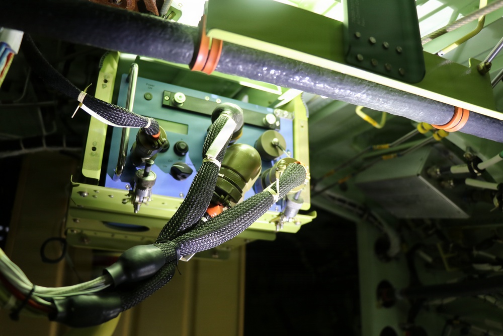 The image shows the interior of what appears to be a military or aerospace vehicle, featuring a complex array of wiring, cables, and electronic equipment mounted on a green-painted structural frame. Multiple braided cable harnesses and connectors are visible, along with what appears to be an avionics or control unit panel with switches and displays. The scene is illuminated by artificial lighting, highlighting the dense technical infrastructure.
