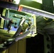 The image shows the interior of what appears to be a military or aerospace vehicle, featuring a complex array of wiring, cables, and electronic equipment mounted on a green-painted structural frame. Multiple braided cable harnesses and connectors are visible, along with what appears to be an avionics or control unit panel with switches and displays. The scene is illuminated by artificial lighting, highlighting the dense technical infrastructure.