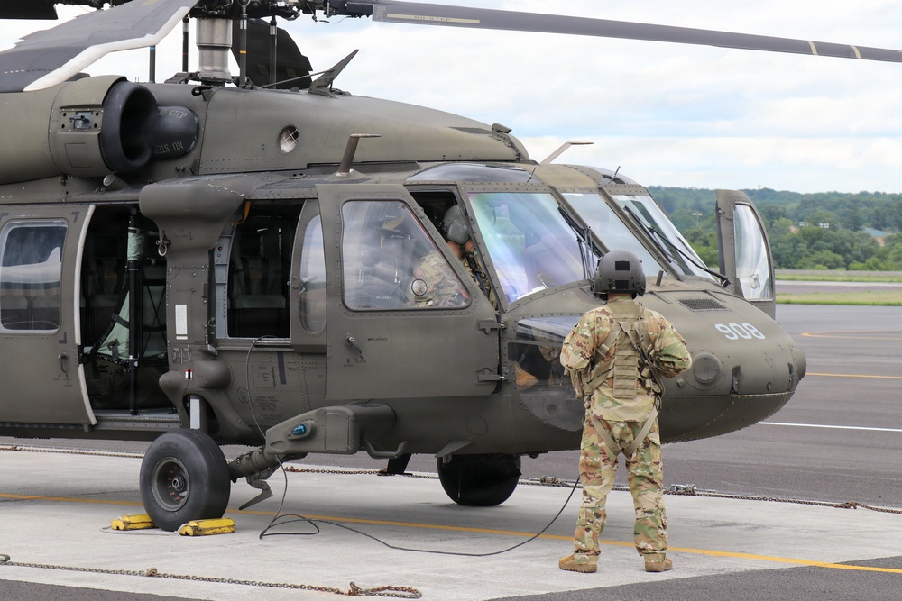 A military helicopter, identified as a UH-60 Black Hawk (tail number 908), is parked on a tarmac with ground crew present. A soldier in OCP (Operational Camouflage Pattern) uniform and flight helmet stands facing the aircraft, holding what appears to be a communication cord or ground power cable. At least one crew member is visible inside the cockpit, suggesting pre-flight or post-flight operations.