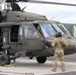 A military helicopter, identified as a UH-60 Black Hawk (tail number 908), is parked on a tarmac with ground crew present. A soldier in OCP (Operational Camouflage Pattern) uniform and flight helmet stands facing the aircraft, holding what appears to be a communication cord or ground power cable. At least one crew member is visible inside the cockpit, suggesting pre-flight or post-flight operations.