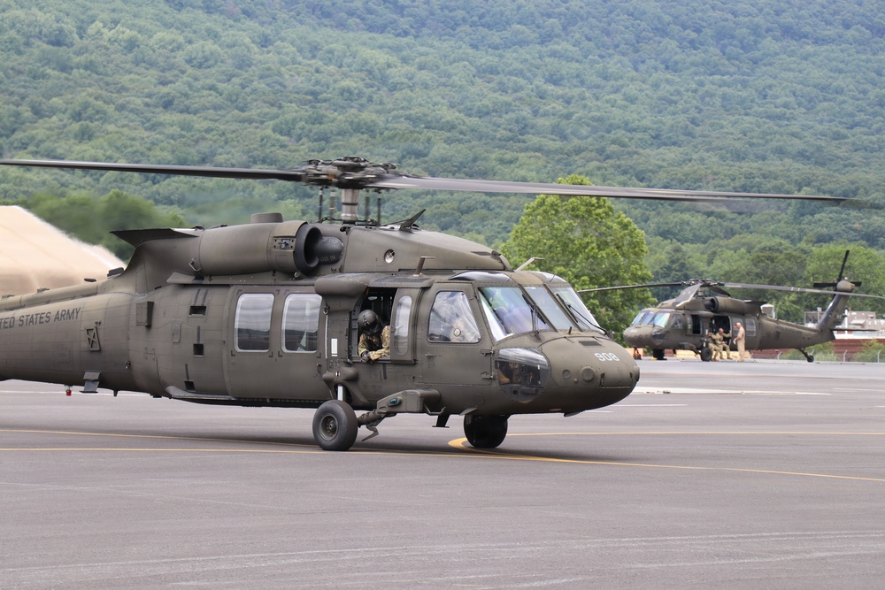 The image shows at least two United States Army UH-60 Black Hawk helicopters on a tarmac or airfield, with wooded hills visible in the background. The foreground helicopter bears the text 'UNITED STATES ARMY' on its fuselage and the tail number '508'. Personnel in military gear are visible both inside and around the aircraft.