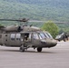The image shows at least two United States Army UH-60 Black Hawk helicopters on a tarmac or airfield, with wooded hills visible in the background. The foreground helicopter bears the text 'UNITED STATES ARMY' on its fuselage and the tail number '508'. Personnel in military gear are visible both inside and around the aircraft.