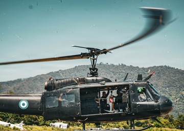 A dark military helicopter, identifiable as a Bell UH-1 'Huey' type, is hovering or just lifting off from a grass airfield with its side door open and crew visible inside. The aircraft bears a roundel insignia featuring a white circle with a blue dot, consistent with certain national air forces. In the background, derelict aircraft frames are visible alongside a small building and a forested hillside.