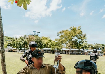 A tactical combat casualty care (TCCC) training exercise is taking place on a grassy field at what appears to be a military installation. A person in multicam uniform is administering an IV drip to a simulated casualty lying on the ground, while personnel in dark digital camouflage uniforms with 'TRIDENTE' patches observe and assist. Additional uniformed personnel with helmets and tactical gear are visible in the background.