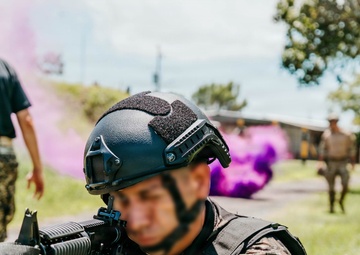 A member of the Marina Nacional (National Navy) wearing tactical gear and a ballistic helmet is aiming a rifle during what appears to be a military training exercise. Purple smoke is visible in the background, commonly used as a training or signaling device. Additional personnel in military attire are visible in the background.