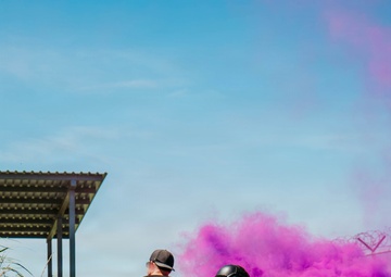 Two individuals are positioned near a deployed purple/magenta smoke grenade on a dirt and gravel surface. One person wears civilian-style black clothing and a cap, while the other is dressed in camouflage uniform with a tactical helmet and backpack. The scene appears to depict a training exercise or tactical drill, with a covered structure and vegetation visible in the background.