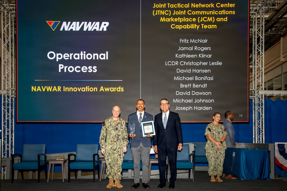 This image captures a NAVWAR Innovation Awards ceremony recognizing the Operational Process category. Three individuals are posed in the foreground — a military officer in Navy Working Uniform, a civilian award recipient holding a plaque and certificate, and another civilian presenter. A large projection screen displays the award category and lists the names of the Joint Tactical Network Center (JTNC) Joint Communications Marketplace (JCM) and Capability Team honorees.