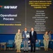This image captures a NAVWAR Innovation Awards ceremony recognizing the Operational Process category. Three individuals are posed in the foreground — a military officer in Navy Working Uniform, a civilian award recipient holding a plaque and certificate, and another civilian presenter. A large projection screen displays the award category and lists the names of the Joint Tactical Network Center (JTNC) Joint Communications Marketplace (JCM) and Capability Team honorees.