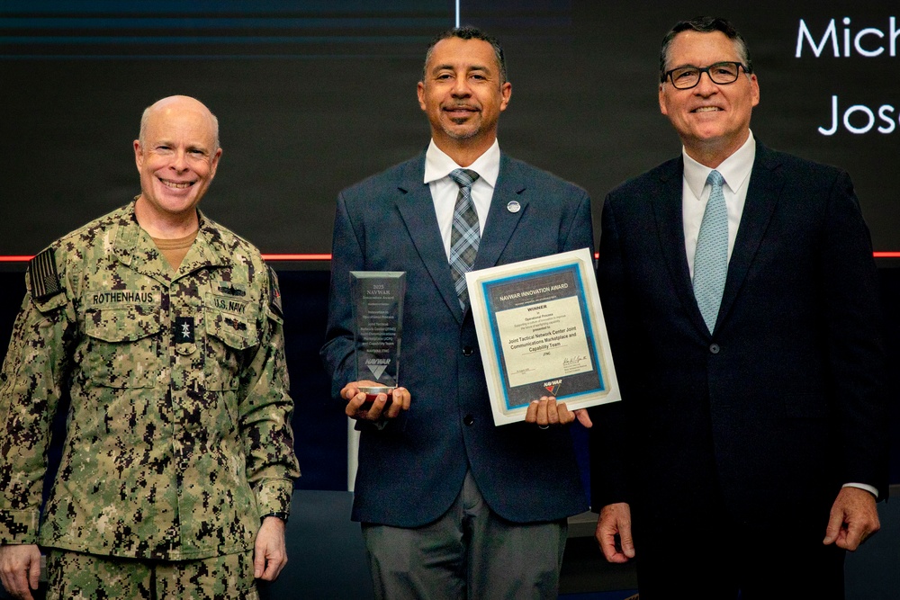 Three men pose together at what appears to be a NAVWAR awards ceremony. The center individual is holding a glass trophy and a framed NAVWAR Innovation Award certificate. The man on the left is a U.S. Navy officer in camouflage uniform with the name tag 'ROTHENHAUS', while the man on the right is in civilian business attire.