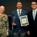 Three men pose together at what appears to be a NAVWAR awards ceremony. The center individual is holding a glass trophy and a framed NAVWAR Innovation Award certificate. The man on the left is a U.S. Navy officer in camouflage uniform with the name tag 'ROTHENHAUS', while the man on the right is in civilian business attire.