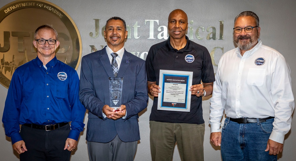 Four individuals are posing for a formal group photo in front of a 'Joint Tactical Network Center' (JTNC) wall sign with a Department of Defense seal. Two of the individuals are holding awards: one holds a glass/crystal trophy and another holds a framed 'NAVWAR Innovation Award' certificate. All four individuals are wearing JTNC logo badges or shirts, indicating organizational affiliation.