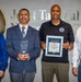 Four individuals are posing for a formal group photo in front of a 'Joint Tactical Network Center' (JTNC) wall sign with a Department of Defense seal. Two of the individuals are holding awards: one holds a glass/crystal trophy and another holds a framed 'NAVWAR Innovation Award' certificate. All four individuals are wearing JTNC logo badges or shirts, indicating organizational affiliation.