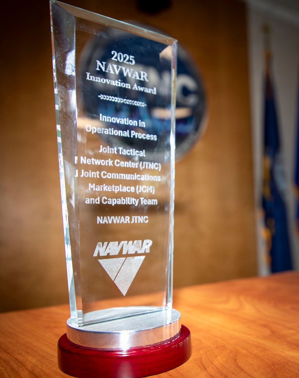 The image shows a crystal glass award trophy presented by NAVWAR in 2025, specifically the 'Innovation in Operational Process' category. The award was given to the Joint Tactical Network Center (JTNC), Joint Communications Marketplace (JCM), and Capability Team under NAVWAR JTNC. The trophy sits on a red and silver base on a wooden surface, with a blurred NAVWAR seal and a blue flag visible in the background.