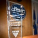 The image shows a crystal glass award trophy presented by NAVWAR in 2025, specifically the 'Innovation in Operational Process' category. The award was given to the Joint Tactical Network Center (JTNC), Joint Communications Marketplace (JCM), and Capability Team under NAVWAR JTNC. The trophy sits on a red and silver base on a wooden surface, with a blurred NAVWAR seal and a blue flag visible in the background.