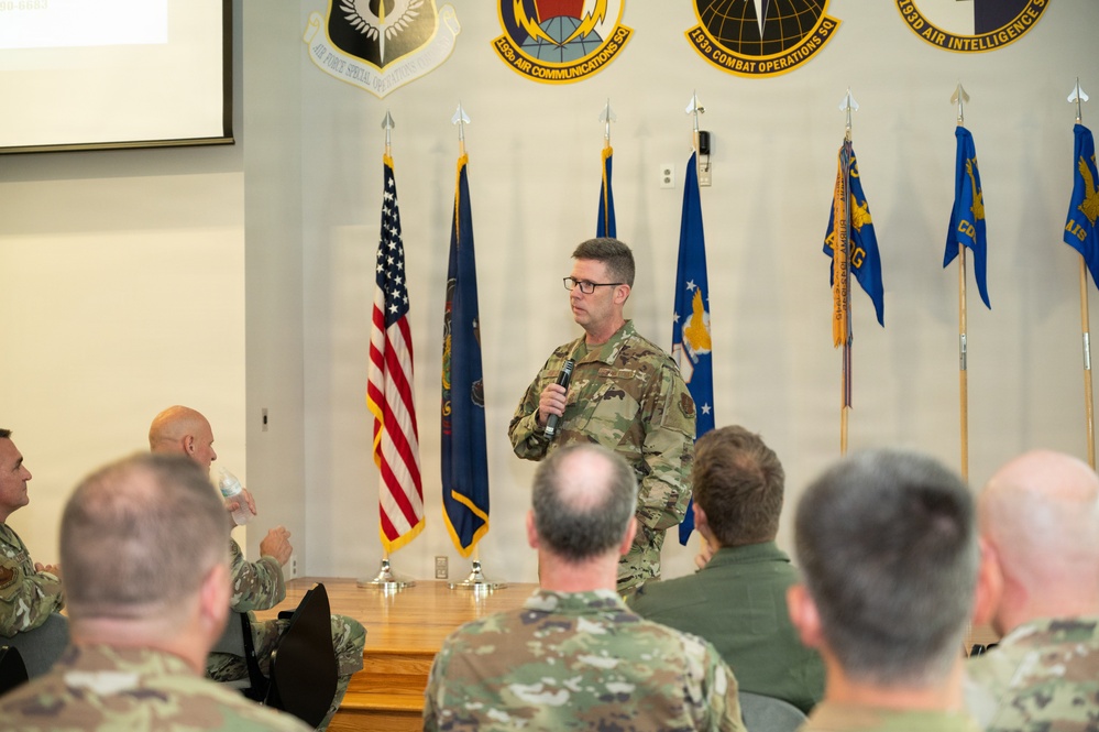 A military officer in OCP (Operational Camouflage Pattern) uniform is addressing an audience of fellow service members in what appears to be a briefing room or conference hall. The speaker is holding a microphone and standing before a display of military unit flags and emblems. The setting appears to be a formal military briefing or ceremony at an Air Force installation.