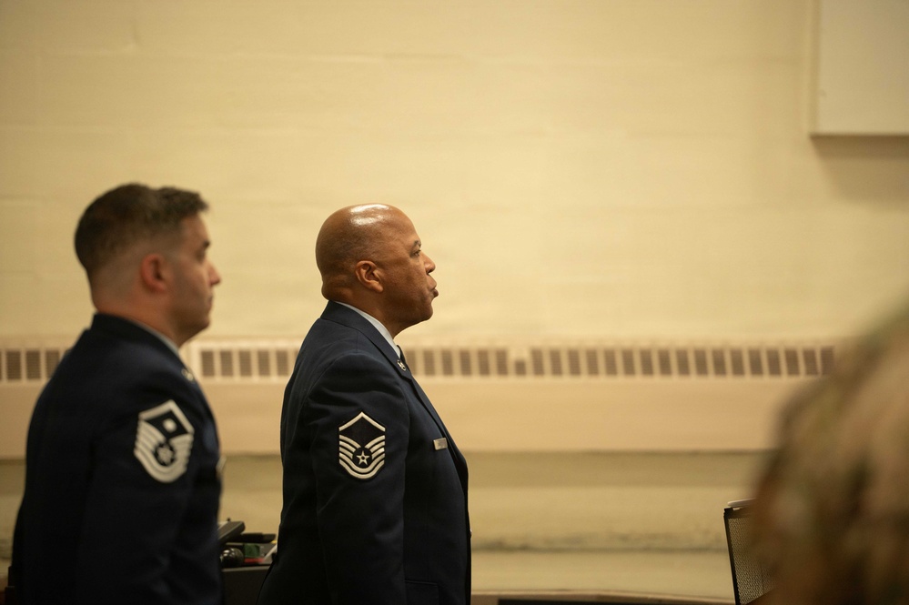 Two U.S. Air Force personnel in dress blue uniforms are standing at attention in what appears to be an indoor military ceremony or formal event. Both individuals display Master Sergeant rank insignia on their sleeves. The image is taken from a side profile angle in a room with beige/tan walls.