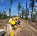 A crew of wildland firefighters in standard yellow Nomex gear and hard hats are advancing in a single-file line along a freshly bulldozed fire line through a conifer forest. The lead firefighter is carrying a coiled fire hose, while others carry hand tools such as pulaskis and shovels. Smoke is faintly visible in the background, suggesting an active or nearby wildfire.
