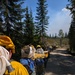 A line of wildland firefighters in yellow Nomex fire-resistant shirts and yellow hard hats are walking along a dirt forest road. They are carrying coiled fire hoses on their backs and collectively transporting what appears to be a long pole or hose lay along the road. Smoke is visible in the sky in the background, indicating an active fire in the vicinity.