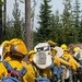 A line of wildland firefighters, viewed from behind, are assembled in a forested area carrying fire hoses and equipment. They are wearing standard wildland firefighting gear including yellow Nomex shirts, green pants, yellow hard hats, and blue NFES (National Fire Equipment System) backpacks. The setting appears to be an active or recently burned forest area during daytime.