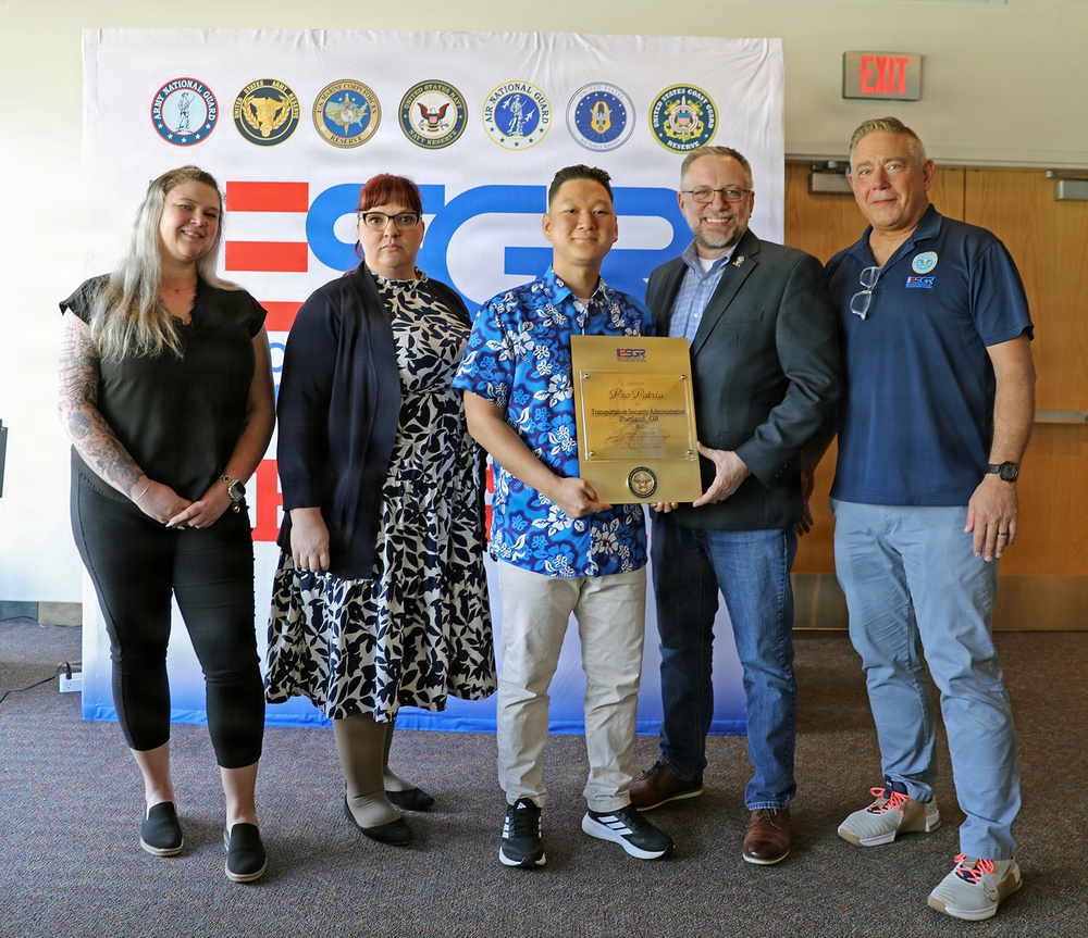 Five individuals are posing for a group photo in front of an ESGR (Employer Support of the Guard and Reserve) banner displaying multiple U.S. military branch seals. Two people in the center are jointly holding a gold ESGR 'Pro Patria' award plaque presented to the Transportation Security Administration. The setting appears to be an indoor ceremony or recognition event.