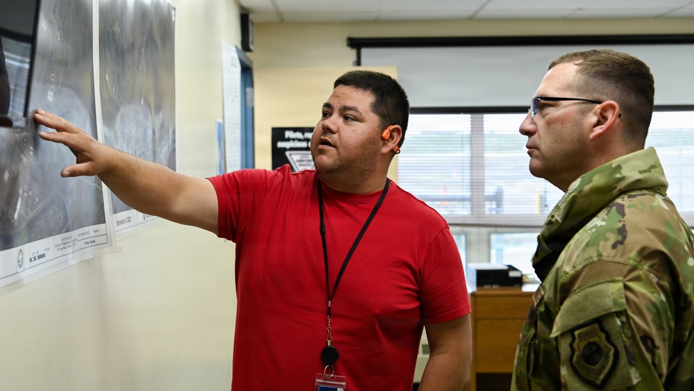 A civilian in a red shirt with a lanyard/badge is briefing a military personnel wearing OCP (Operational Camouflage Pattern) uniform in what appears to be an office or operations center. The civilian is pointing to aerial or satellite imagery maps posted on a wall. The setting suggests a planning or intelligence briefing session.