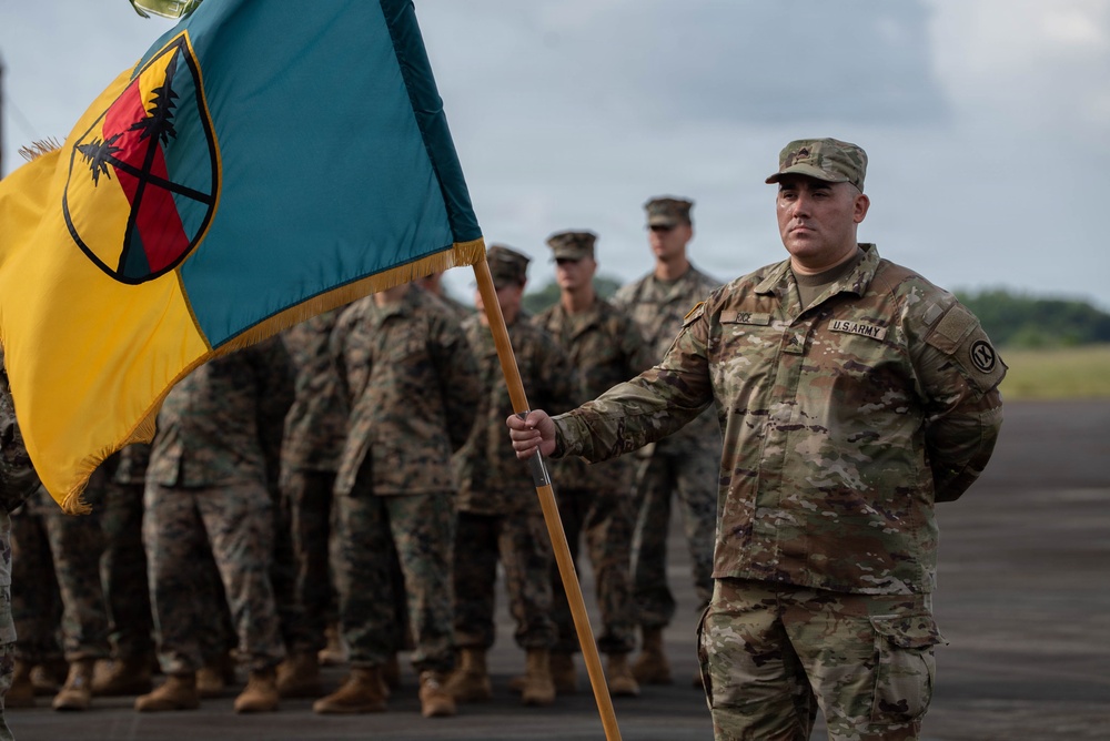 A U.S. Army soldier stands at attention holding a military unit guidon/flag during what appears to be a formal military ceremony on a tarmac or parade ground. Behind him, a formation of military personnel in camouflage uniforms — appearing to include both U.S. Army and U.S. Marine Corps members — stand at ease. The flag features a teal and yellow design with a circular emblem containing crossed elements and a tree motif.