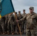 A U.S. Army soldier stands at attention holding a military unit guidon/flag during what appears to be a formal military ceremony on a tarmac or parade ground. Behind him, a formation of military personnel in camouflage uniforms — appearing to include both U.S. Army and U.S. Marine Corps members — stand at ease. The flag features a teal and yellow design with a circular emblem containing crossed elements and a tree motif.