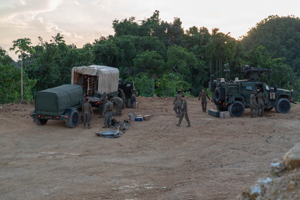 A group of military personnel in camouflage uniforms are conducting field operations on a cleared dirt area surrounded by dense tropical vegetation. Multiple military vehicles are present, including covered cargo trucks and what appears to be an armored tactical vehicle (likely a JLTV or similar platform) equipped with a weapon mount and communications equipment. The scene appears to be either a setup or breakdown of a forward operating position, photographed during dusk or dawn lighting conditions.