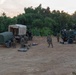 A group of military personnel in camouflage uniforms are conducting field operations on a cleared dirt area surrounded by dense tropical vegetation. Multiple military vehicles are present, including covered cargo trucks and what appears to be an armored tactical vehicle (likely a JLTV or similar platform) equipped with a weapon mount and communications equipment. The scene appears to be either a setup or breakdown of a forward operating position, photographed during dusk or dawn lighting conditions.