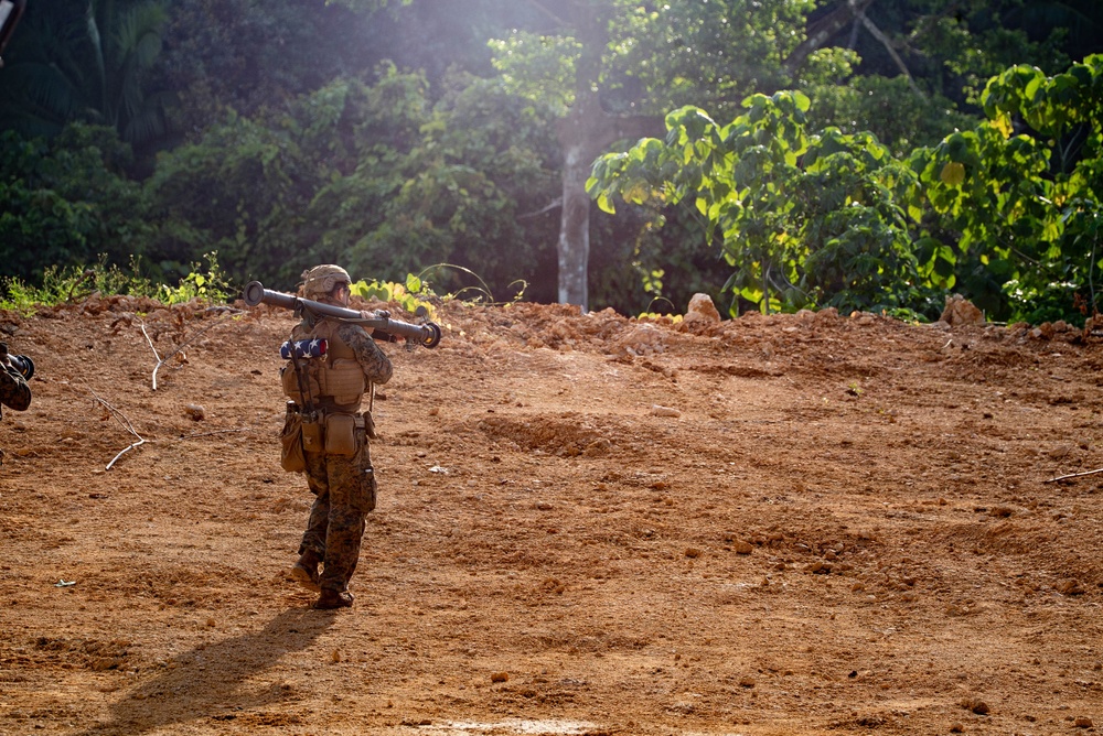 A single military service member in camouflage uniform is carrying a shoulder-launched weapon system across an open dirt clearing. The soldier is facing away from the camera and appears to be moving toward a treeline in a tropical or subtropical environment. An American flag patch is visible on the back of the soldier's pack.