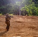 A single military service member in camouflage uniform is carrying a shoulder-launched weapon system across an open dirt clearing. The soldier is facing away from the camera and appears to be moving toward a treeline in a tropical or subtropical environment. An American flag patch is visible on the back of the soldier's pack.