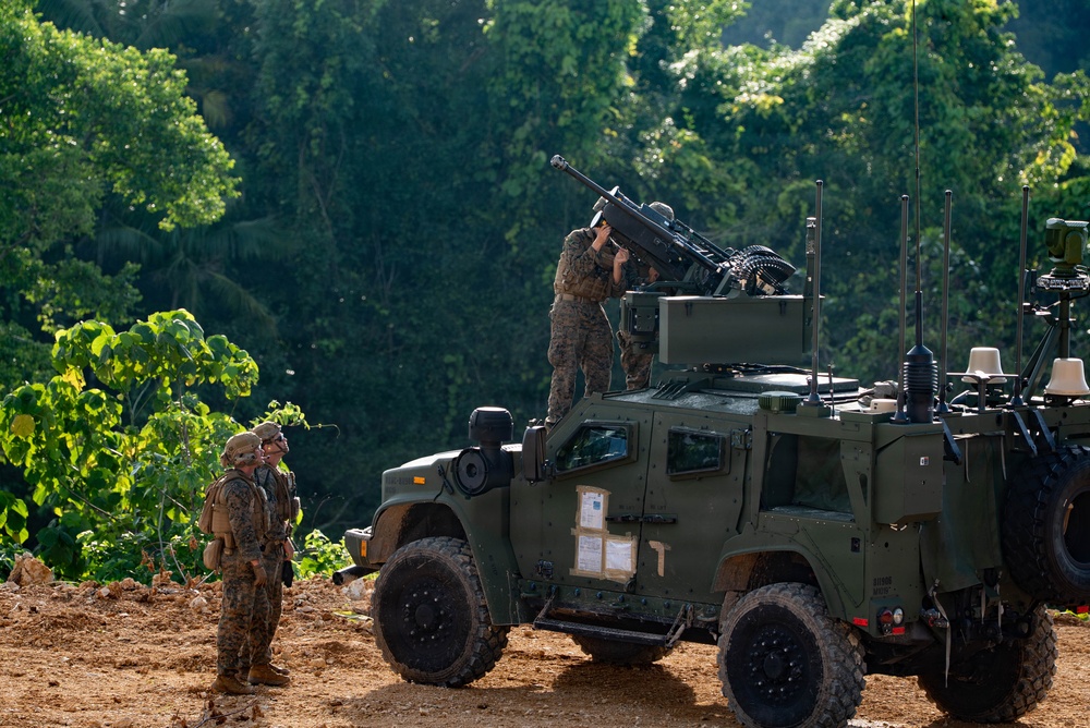 The image shows military personnel in camouflage uniforms operating around a Joint Light Tactical Vehicle (JLTV) mounted with a crew-served weapon, likely an M2 .50 caliber machine gun or Mk 19 grenade launcher. The setting appears to be a tropical or subtropical environment with dense green vegetation in the background. Two soldiers stand to the left observing while one soldier atop the vehicle appears to be configuring or loading the mounted weapon system.