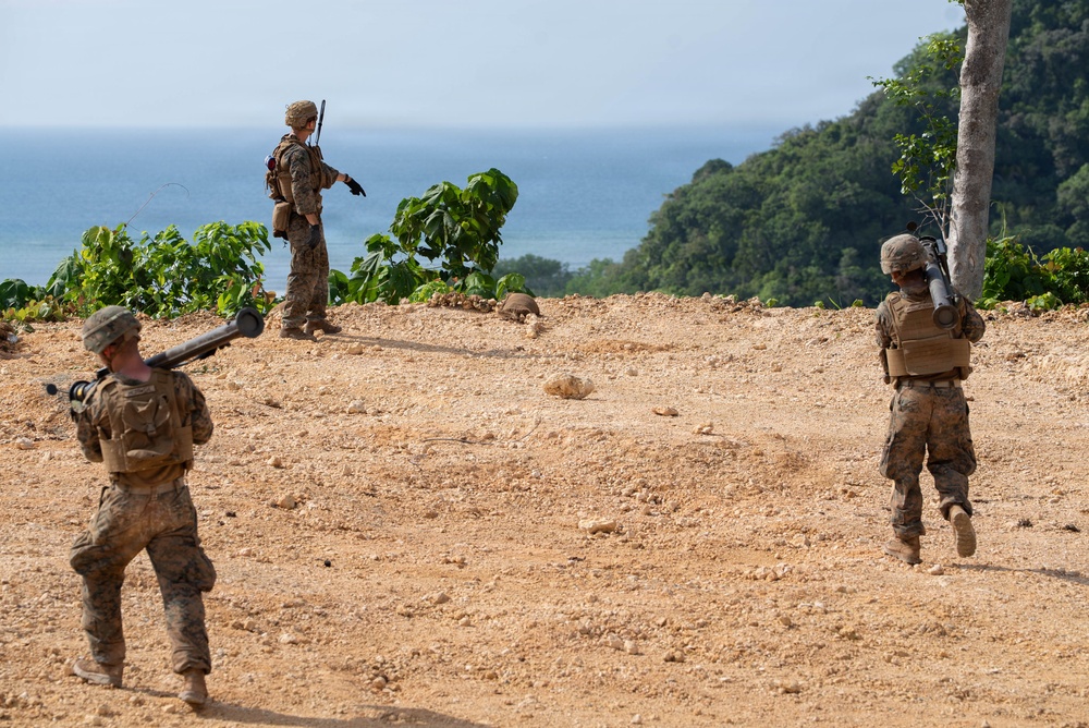 Three military personnel in camouflage uniforms are positioned on a sandy elevated terrain overlooking a body of water. Two of the soldiers are carrying what appear to be shoulder-launched rocket or missile systems. The setting appears to be a coastal tropical environment with dense green vegetation visible in the background.