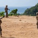 Three military personnel in camouflage uniforms are positioned on a sandy elevated terrain overlooking a body of water. Two of the soldiers are carrying what appear to be shoulder-launched rocket or missile systems. The setting appears to be a coastal tropical environment with dense green vegetation visible in the background.