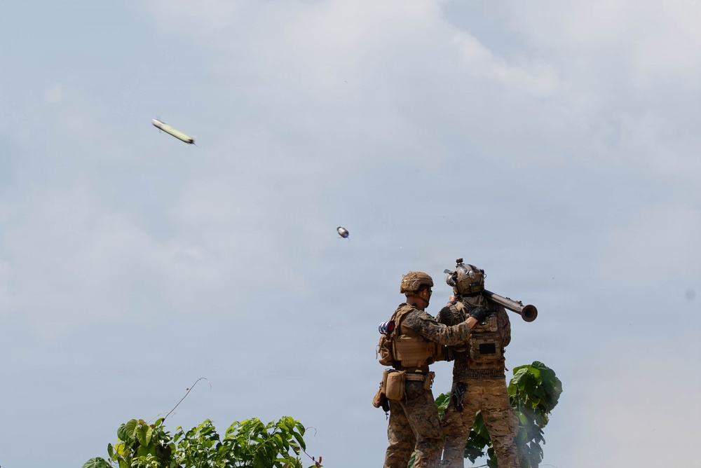Two soldiers in camouflage uniforms and tactical gear are operating what appears to be a man-portable air-defense system (MANPADS) or shoulder-fired rocket launcher. A projectile or missile is visible in flight in the upper portion of the image against a partly cloudy sky. The scene appears to be an active firing exercise or combat operation in a tropical or subtropical environment.