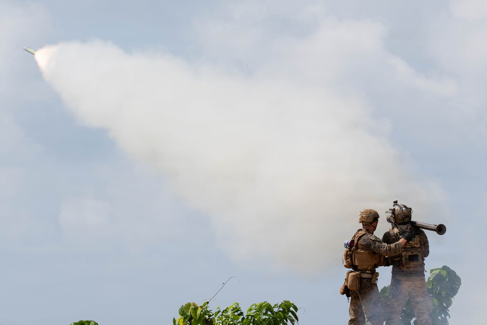 Two soldiers in full combat gear are operating a man-portable surface-to-air missile launcher, with a missile visibly in flight leaving a large white smoke trail across the sky. The soldiers are positioned on elevated ground surrounded by vegetation. The image appears to capture a live-fire military exercise or combat operation.