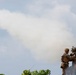Two soldiers in full combat gear are operating a man-portable surface-to-air missile launcher, with a missile visibly in flight leaving a large white smoke trail across the sky. The soldiers are positioned on elevated ground surrounded by vegetation. The image appears to capture a live-fire military exercise or combat operation.