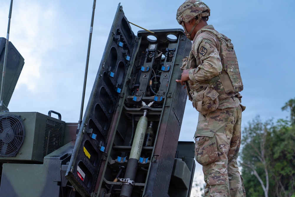 A U.S. Army soldier in OCP (Operational Camouflage Pattern) uniform is working on what appears to be a Patriot missile defense system launcher unit. The soldier is inspecting or maintaining the open access panel of the launcher, which reveals internal components including wiring, connectors, and a visible missile canister. The setting appears to be an outdoor field environment under a partly cloudy sky.