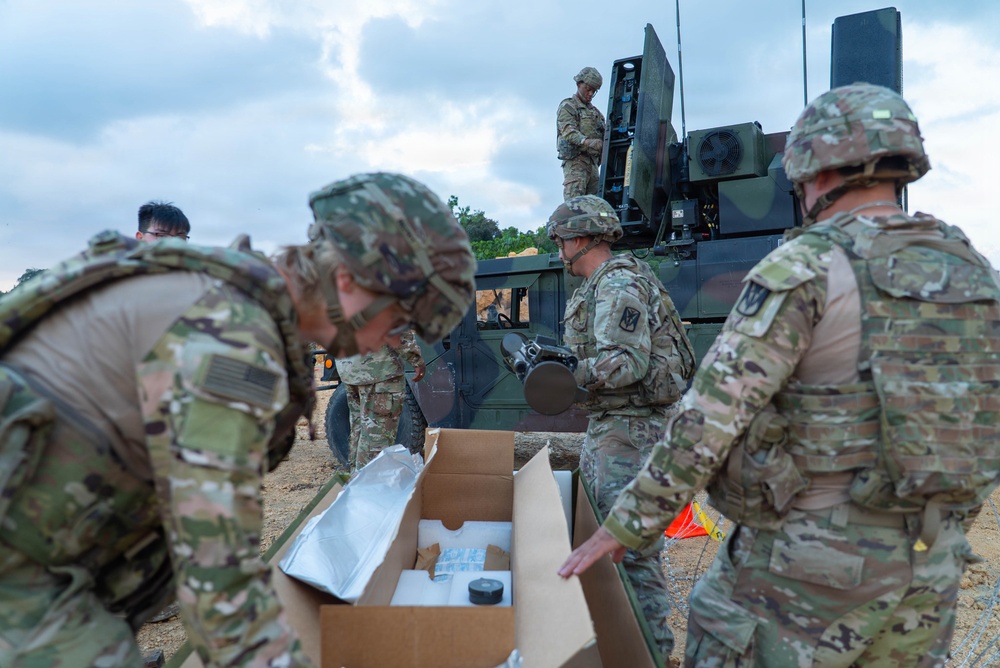 A group of soldiers in MultiCam/OCP camouflage uniforms are working around a military vehicle, appearing to unpack and handle equipment components. One soldier is positioned on top of the vehicle while others work at ground level with open cardboard boxes containing what appear to be equipment parts. The setting is an outdoor field environment with sandy/dirt terrain under a partly cloudy sky.