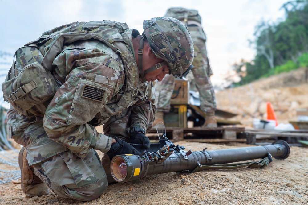 A U.S. Army soldier in MultiCam/OCP camouflage uniform is kneeling on sandy ground, closely inspecting or preparing a shoulder-fired weapon system. A second soldier is visible in the background performing a similar task. The setting appears to be a military training range, indicated by the safety cones and wooden pallets visible in the background.
