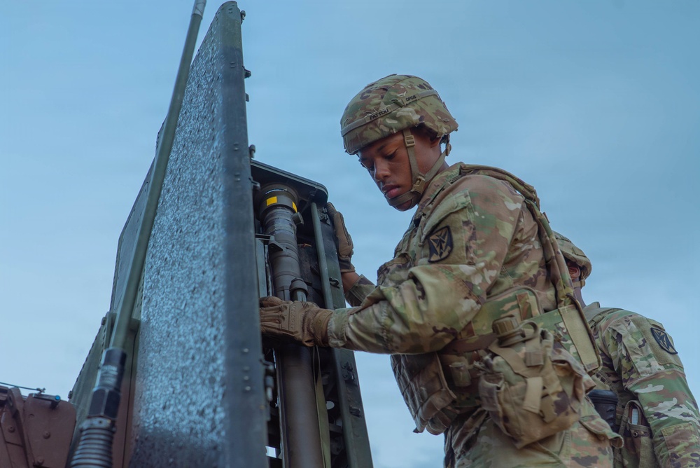 A U.S. Army soldier in full combat gear is working on what appears to be a Patriot missile system or similar military air defense equipment. The soldier is wearing MultiCam (OCP) camouflage uniform with a combat helmet and tactical vest. A second soldier is partially visible in the background, suggesting a team operation.
