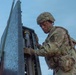 A U.S. Army soldier in full combat gear is working on what appears to be a Patriot missile system or similar military air defense equipment. The soldier is wearing MultiCam (OCP) camouflage uniform with a combat helmet and tactical vest. A second soldier is partially visible in the background, suggesting a team operation.