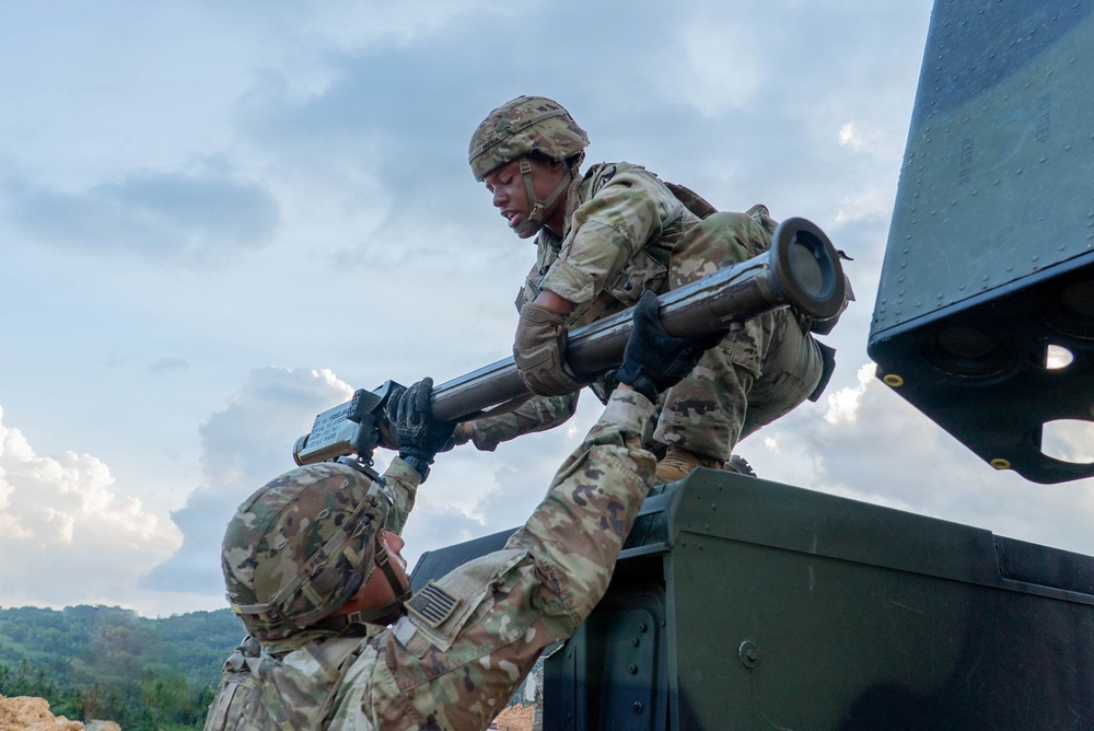 Two U.S. Army soldiers in OCP (Operational Camouflage Pattern) uniforms are handling what appears to be a Stinger man-portable air-defense missile system (MANPADS) on or near a military vehicle. One soldier is positioned on top of the vehicle while the other assists from below, passing or receiving the weapon system. The setting appears to be an outdoor training or operational environment with hilly, vegetated terrain visible in the background.