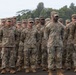 A formation of U.S. Army soldiers standing at attention on what appears to be a tarmac or airfield. The soldiers are wearing Operational Camouflage Pattern (OCP) uniforms with patrol caps and tan combat boots. The setting appears to be a tropical or subtropical location based on the lush green vegetation visible in the background.