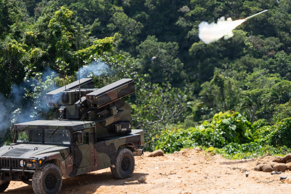 A military HMMWV (Humvee) mounted with a surface-to-air missile launcher system is actively firing a missile. The vehicle is positioned on a dirt clearing surrounded by dense tropical vegetation. A missile is visibly in flight with a white smoke trail against a backdrop of green jungle canopy.