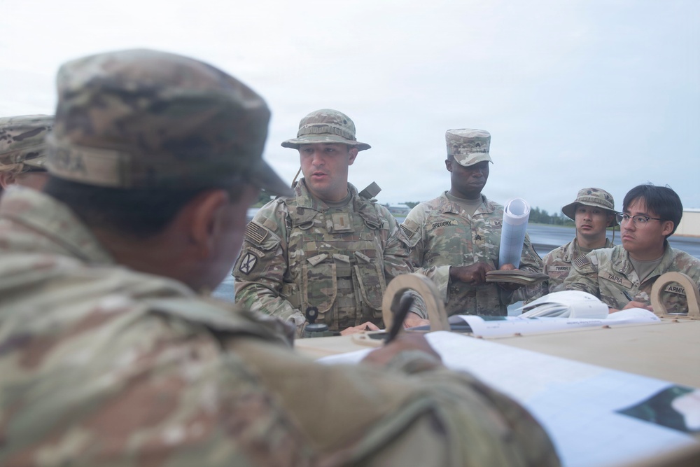 A group of U.S. Army soldiers in OCP (Operational Camouflage Pattern) uniforms are gathered around a table outdoors, appearing to review maps or documents during what looks like a planning or briefing session. One soldier in the foreground is writing or pointing at documents on the table while others observe and listen. Name tapes visible include 'GREGORY', 'TORRES', and 'TARA'.