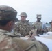 A group of U.S. Army soldiers in OCP (Operational Camouflage Pattern) uniforms are gathered around a table outdoors, appearing to review maps or documents during what looks like a planning or briefing session. One soldier in the foreground is writing or pointing at documents on the table while others observe and listen. Name tapes visible include 'GREGORY', 'TORRES', and 'TARA'.