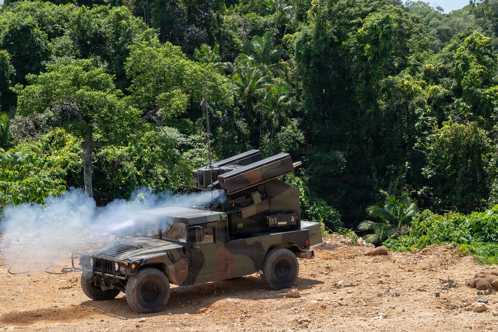 A military HMMWV (Humvee) mounted with a missile or rocket launcher system is captured mid-firing, with visible exhaust smoke emanating from the launcher. The vehicle is positioned on a dirt clearing surrounded by dense tropical or subtropical jungle vegetation. The image appears to document a live-fire exercise or weapons demonstration.