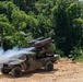 A military HMMWV (Humvee) mounted with a missile or rocket launcher system is captured mid-firing, with visible exhaust smoke emanating from the launcher. The vehicle is positioned on a dirt clearing surrounded by dense tropical or subtropical jungle vegetation. The image appears to document a live-fire exercise or weapons demonstration.